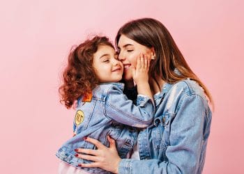 Relaxed Preteen Girl Embracing With Mother. Winsome Young Mom Kissing Daughter On Pink Background