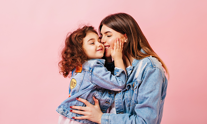 Relaxed Preteen Girl Embracing With Mother. Winsome Young Mom Kissing Daughter On Pink Background